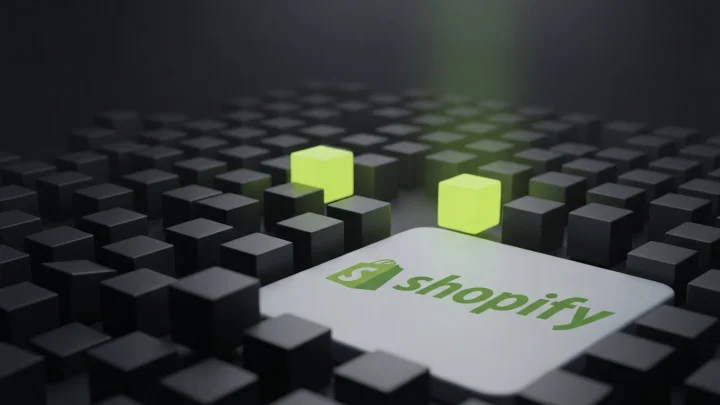 A wide field of dark cubes receding into shadow with a narrow lime light picking out exactly three, and the Shopify logo on a plate in the foreground.